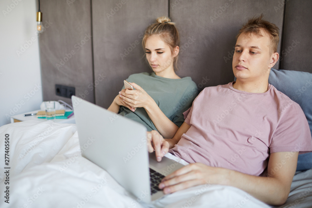 Young man with laptop networking in bed while his wife scrolling in smartphone Young man with laptop networking in bed while his wife scrolling in smartphone