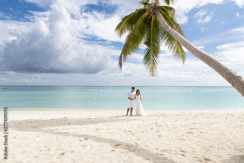 Newlyweds hug under a palm tree on a gorgeous beach with white sand and turquoise water.