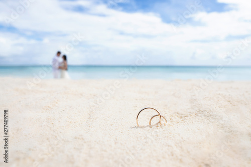 Wedding rings on the white sand. In the background the newlyweds and the ocean.