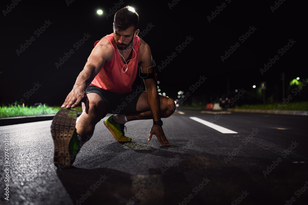 Man preparing to run through the city at night, he stretching his leg muscles. - obrazy, fototapety, plakaty 