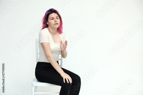 Portrait below the knee of a young beautiful girl teenager in a white T-shirt and black jeans with beautiful purple hair sitting on a white background in the studio. Says, shows hands with emotions.
