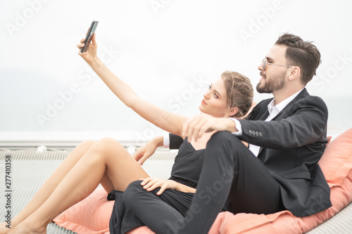 Happy lovely young woman with her handsome boyfriend in suit together taking a selfie on the yacht, smiling and looking at camera. Relax time.