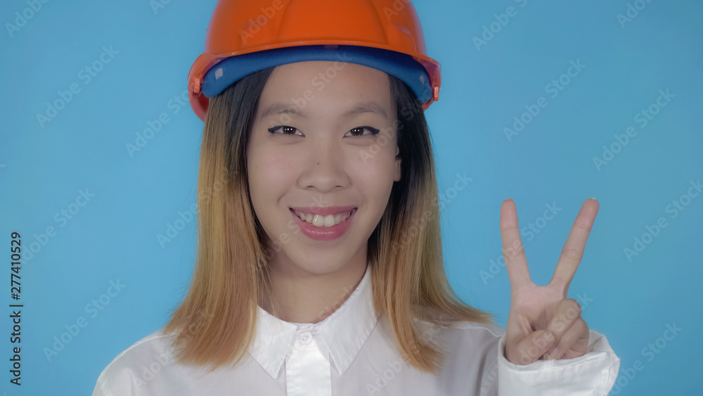 close up portrait young asian architect wearing orange hard hat shows symbol victory on blue background in studio. attractive millennial girl looking at the camera close up portrait young asian architect wearing orange hard hat shows symbol victory on blue background in studio. attractive millennial girl looking at the camera