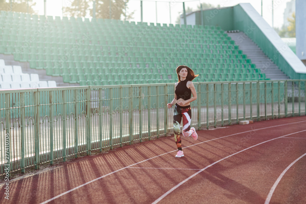 Sport. Athletic young brunette woman in pink sneakers, leggings and top run on running track stadium at sunset. her hair is developing. Concept run. concept of a healthy lifestyle.