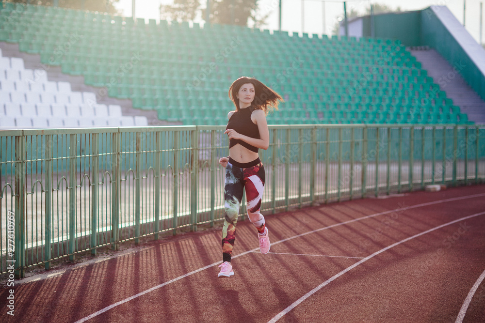 Sport. Athletic young brunette woman in pink sneakers, leggings and top run on running track stadium at sunset. her hair is developing. Concept run. concept of a healthy lifestyle.