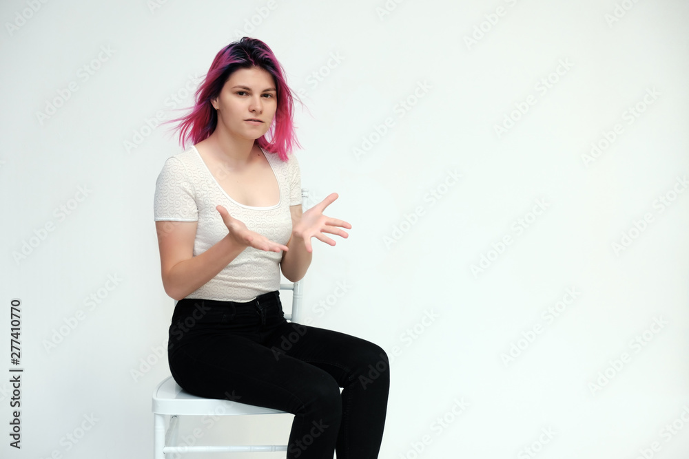 Portrait below the knee of a young beautiful girl teenager in a white T-shirt and black jeans with beautiful purple hair sitting on a white background in the studio. Says, shows hands with emotions. Portrait below the knee of a young beautiful girl teenager in a white T-shirt and black jeans with beautiful purple hair sitting on a white background in the studio. Says, shows hands with emotions.