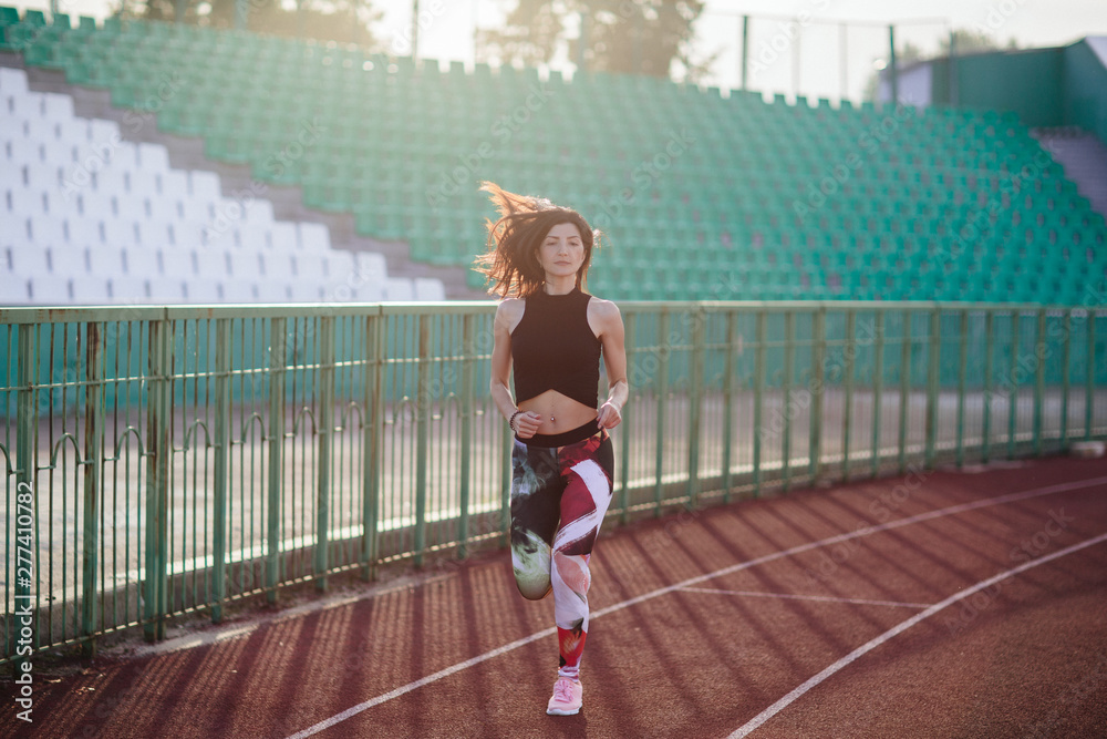 Sport. Athletic young brunette woman in pink sneakers, leggings and top run on running track stadium at sunset. her hair is developing. Concept run. concept of a healthy lifestyle.