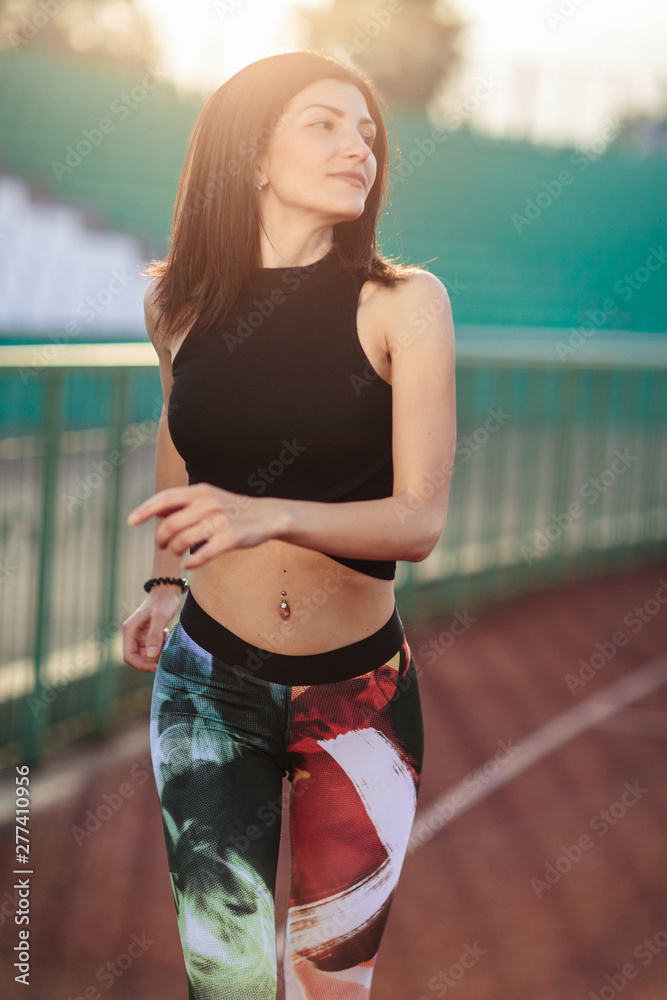Portrait of Athletic young brunette woman in pink sneakers, leggings and top run on running track stadium at sunset. her hair is developing. Concept run. concept of a healthy lifestyle.