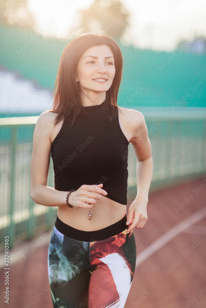 Portrait of Athletic young brunette woman in pink sneakers, leggings and top run on running track stadium at sunset. her hair is developing. Concept run. concept of a healthy lifestyle.