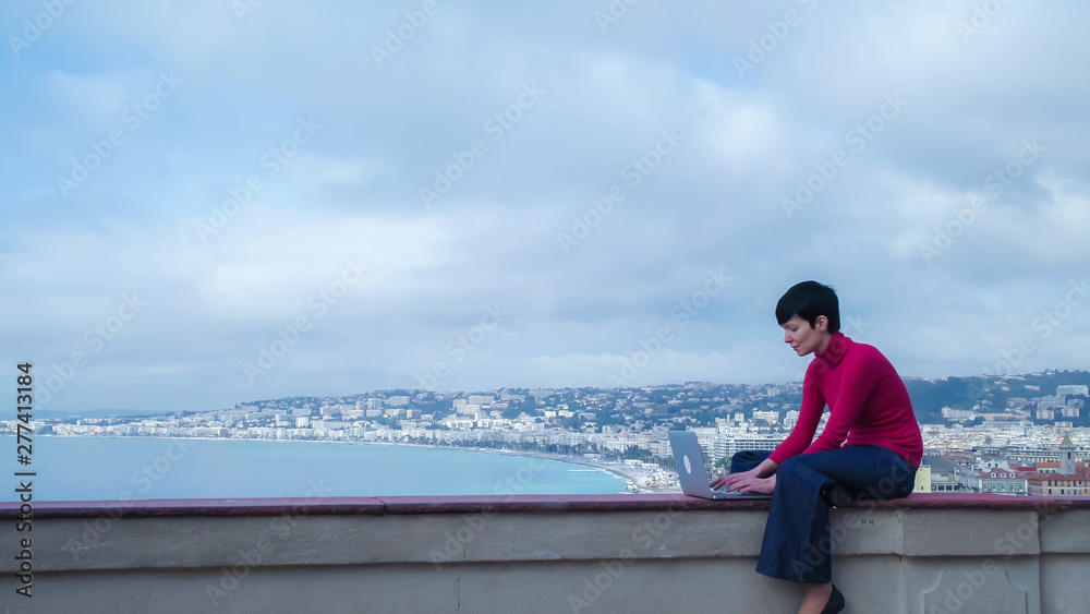 Female sitting in front of sea and slouch. Caucasian model wearing in jeans and red pullover using laptop outdoor. On the background seascape with sea beach and seafront in the marine city.