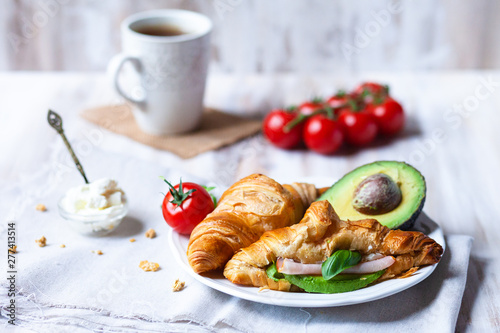 Delicious breakfast french style at home: light background, white plate with fresh croissants, small bowls of granola and cream cheese, cup of tea, avocado, tomatoes, basil. Served on beige napkins