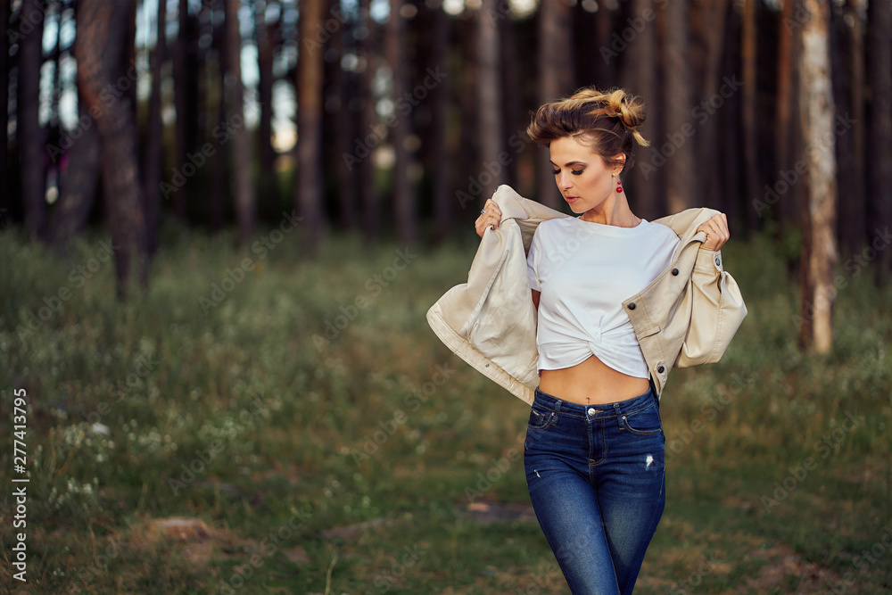 girl in jeans and a white t-shirt. standing in a light jacket against a background of pine forest at sunset