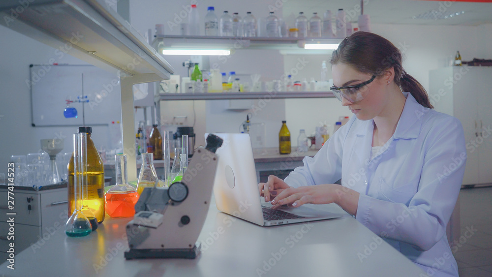 Fototapeta premium Portrait adult caucasian doctor working with laptop in laboratory. Chemist engineer or lab technician entering data on laptop. Professional woman working in the evening alone.