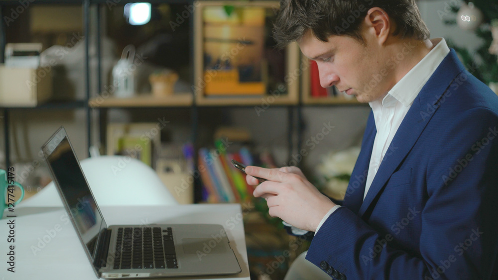 Handsome man sitting in room with bookshelves on the desk laptop. Elegant fashionable man wearing in casual blue suit texting message on smartphone.