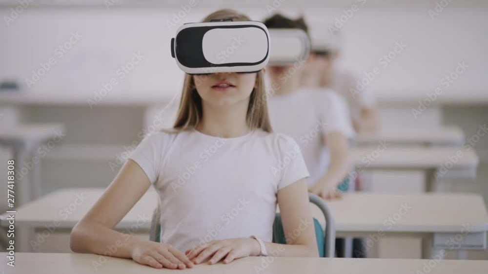 Surprised curious school kids using VR headsets watching 3D cartoons during a lesson. Focus on an amazed young girl in white sitting by desk. Surprised curious school kids using VR headsets watching 3D cartoons during a lesson. Focus on an amazed young girl in white sitting by desk.