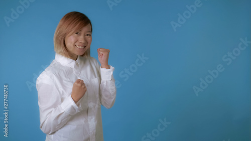 portrait young asian female posing happy dancing on blue background in studio. attractive korean woman with blond hair wearing white casual shirt looking at the camera. © vadosloginov