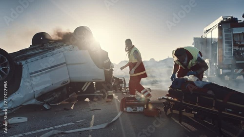 On the Car Crash Traffic Accident Scene: Paramedics Giving First Aid Oxygen Mask to Female Victim of the Accident. Firefighters Extinguish Fire and Use Hydraulic Cutter to Free Other Passengers