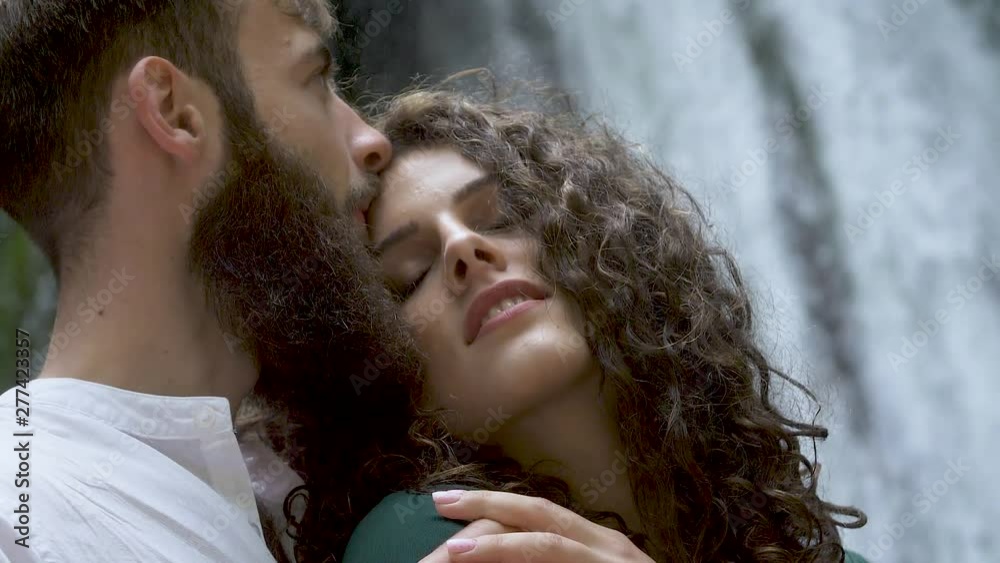 attractive man with a beard hugging a woman with curly hair from behind who leaned on his chest and kisses her on the forehead against the backdrop of a waterfall in a mountain forest Slow motion