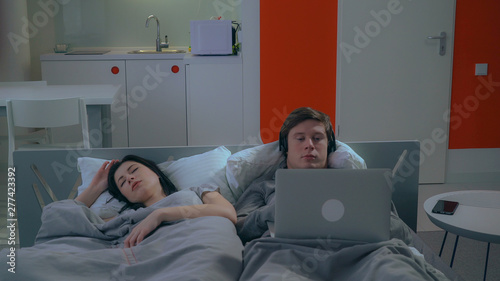 family in bed in the morning. Young woman sleeping . Caucasian man lying with computer and headphone. Guy listen music or watch movie in apartment. photo