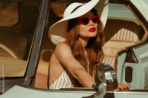 Street fashion portrait of young elegant luxury lady wearing white sunglasses, wide brim hat, striped linen jumpsuit, posing in the retro car. Copy, empty space for text