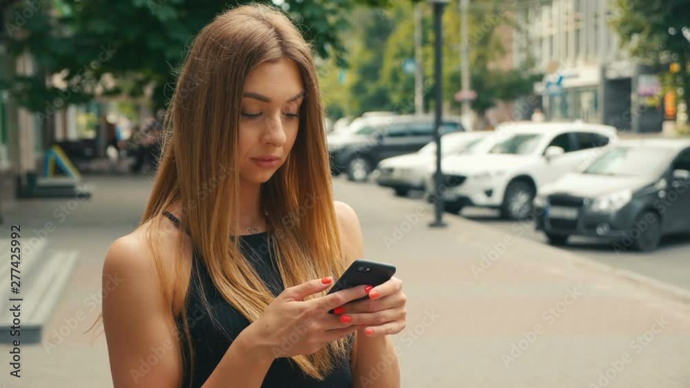 Pretty young woman typing on mobile phone with people walking in street at background, Brown haired woman looking at smartphone in hand
