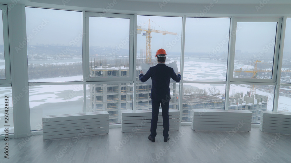elegant engineer or architect, or manager in real estate company. businessman enters the room. Rear back view male holding architectural plan looking through the big windows on the construction site elegant engineer or architect, or manager in real estate company. businessman enters the room. Rear back view male holding architectural plan looking through the big windows on the construction site