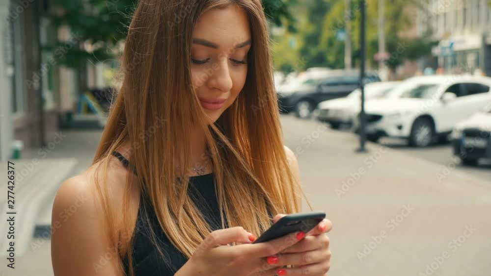 Pretty young woman typing on mobile phone with people walking in street at background, Brown haired woman looking at smartphone in hand