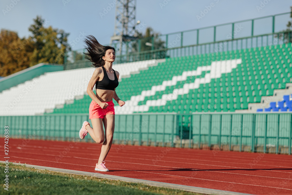 Young fitness brunette woman in shorts and top running on a stadium track. Athlete girl doing exercises on the training at stadium. Healthy active lifestyle.