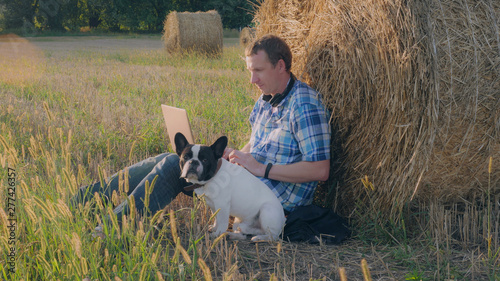 Caucasian man with french bulldog sitting on the field. Male wearing casual shirt and jeans using laptop. © vadosloginov