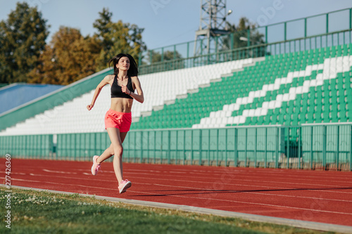 Young fitness brunette woman in shorts and top running on a stadium track. Athlete girl doing exercises on the training at stadium. Healthy active lifestyle.