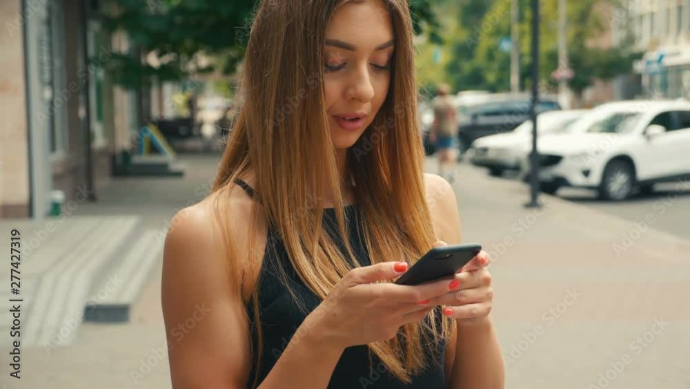 Pretty young woman typing on mobile phone with people walking in street at background, Brown haired woman looking at smartphone in hand