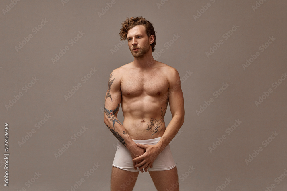 Isolated image of gorgeous handsome man with stubble and curly hairstyle posing naked in studio wearing only white boxer shorts, holding both hands on groin area, having serious facial expression