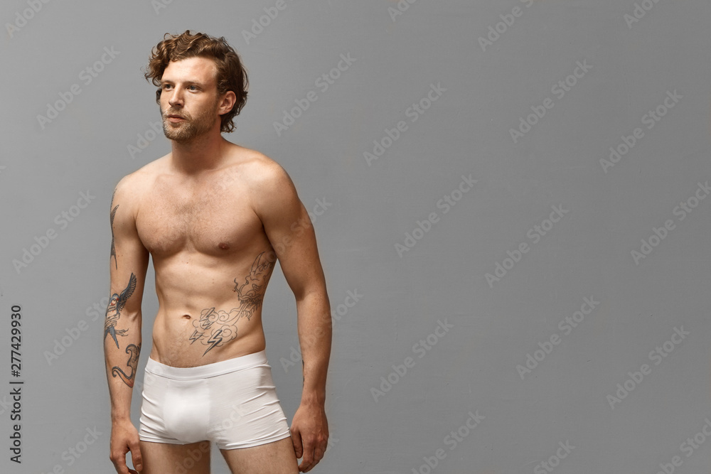 Isolated shot of attractive athletic man with stylish hairdo and tattoos on arm and naked torso wearing only white boxer shorts posing at blank studio wall with copyspace for your advertisement