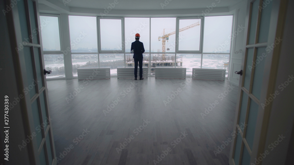 Man wearing in suit and helmet holding documents with architectural plan. rear back view businessman looking through the windows on the construction site with high-rise Man wearing in suit and helmet holding documents with architectural plan. rear back view businessman looking through the windows on the construction site with high-rise