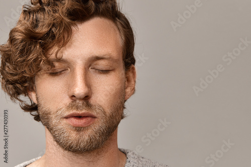 Close up portrait of handsome unshaven young male with stylish hairdo and freckles having peaceful calm facial expression keeping eyes closed, practicing meditation in the morning. Harmony and balance