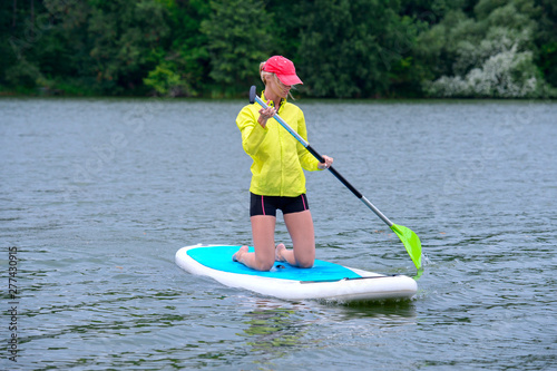 girl in a tracksuit kneels on a surfboard and holds a paddle. Front View Photo