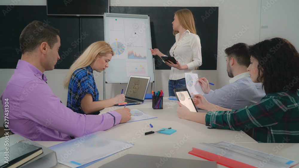 Young woman leader giving presentation standing near flipchart using digital tablet. Staff sitting in boardroom listening about new sales and growth strategy.