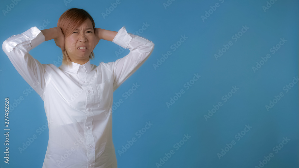 Naklejka premium portrait young asian female hears a terrible sound or scream on blue background in studio. attractive korean woman with blond hair wearing white casual shirt looking at the camera.