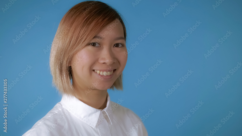 close up face young asian female posing on blue background in studio. attractive korean woman with blond hair wearing white casual shirt looking at the camera laughing close up face young asian female posing on blue background in studio. attractive korean woman with blond hair wearing white casual shirt looking at the camera laughing