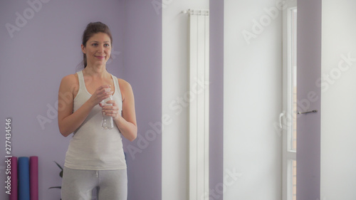 Woman drinks water from a bottle in a gym, happy and smiling.