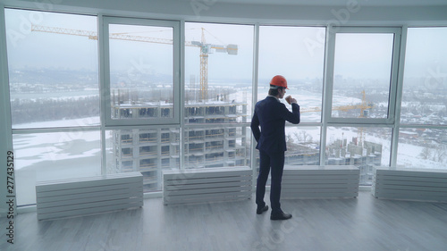 businessman in the white office looking on the street with construction site wearing in elegant suit drinking coffee oversees how workers working. rear back view man with orange hard hat in modern