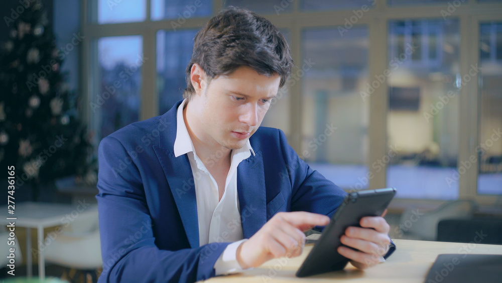 Portrait handsome adult man using touch screen tablet. Caucasian young male surfing internet or chatting in application. Businessman wearing in casual elegant jacket sitting in room with big windows Portrait handsome adult man using touch screen tablet. Caucasian young male surfing internet or chatting in application. Businessman wearing in casual elegant jacket sitting in room with big windows