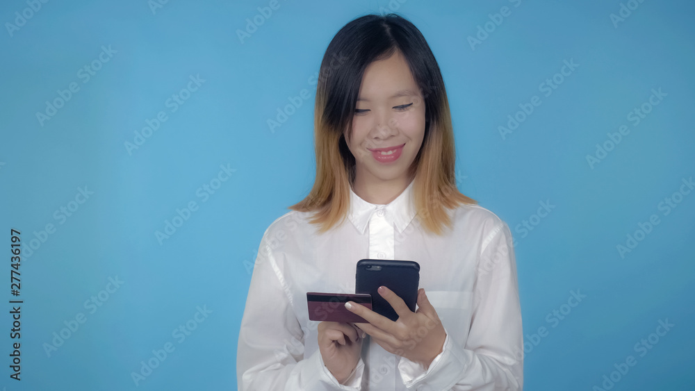 young asian woman using smartphone for buying online or pay order or bills on blue background in studio. attractive millennial girl wearing white casual shirt looking at the camera smiling