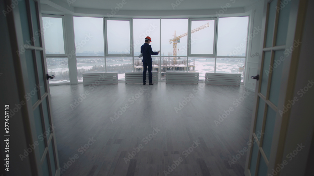 rear back view manager in contemporary company real estate property check documents and looking how growing house on construction site. elegant man in suit and hard hat in white modern develop office.