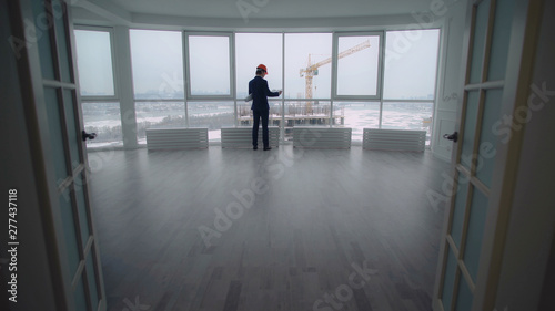 rear back view manager in contemporary company real estate property check documents and looking how growing house on construction site. elegant man in suit and hard hat in white modern develop office. © vadosloginov