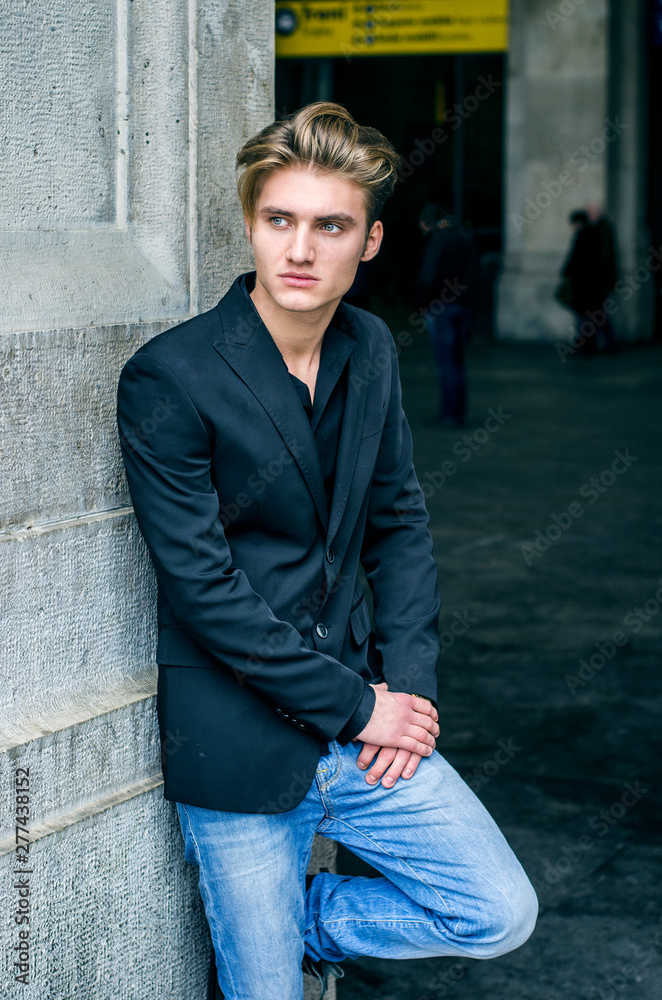 Attractive blue eyed, blond young man leaning with his back against white wall outdoors Attractive blue eyed, blond young man leaning with his back against white wall outdoors