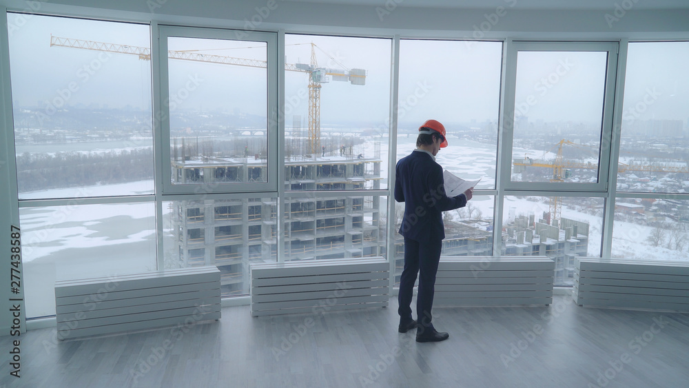 architect holding a design plan or other technical drawing looking on the project . worker in elegant suit and hard hat looking through the windows on the construction site with crane and workers on architect holding a design plan or other technical drawing looking on the project . worker in elegant suit and hard hat looking through the windows on the construction site with crane and workers on