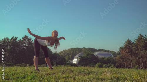 Woman doing stretching exercises in the park with beautiful nature standing on the meadow with green grass and cute yellow flowers. Attractive girl with blond hair strength body before pilates workout