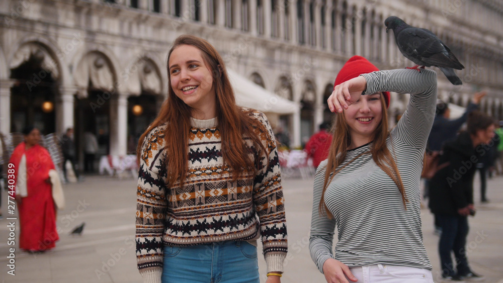 Two young women feed the pigeons from their hands in the city square of Venice.