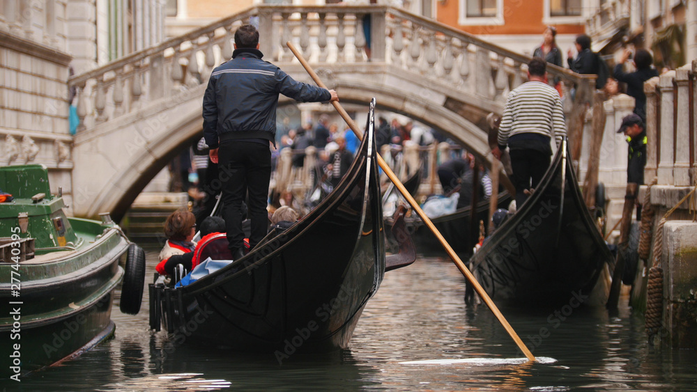 A man stands on a boat with people holding a paddle in his hand - Journey about the canals of Venice